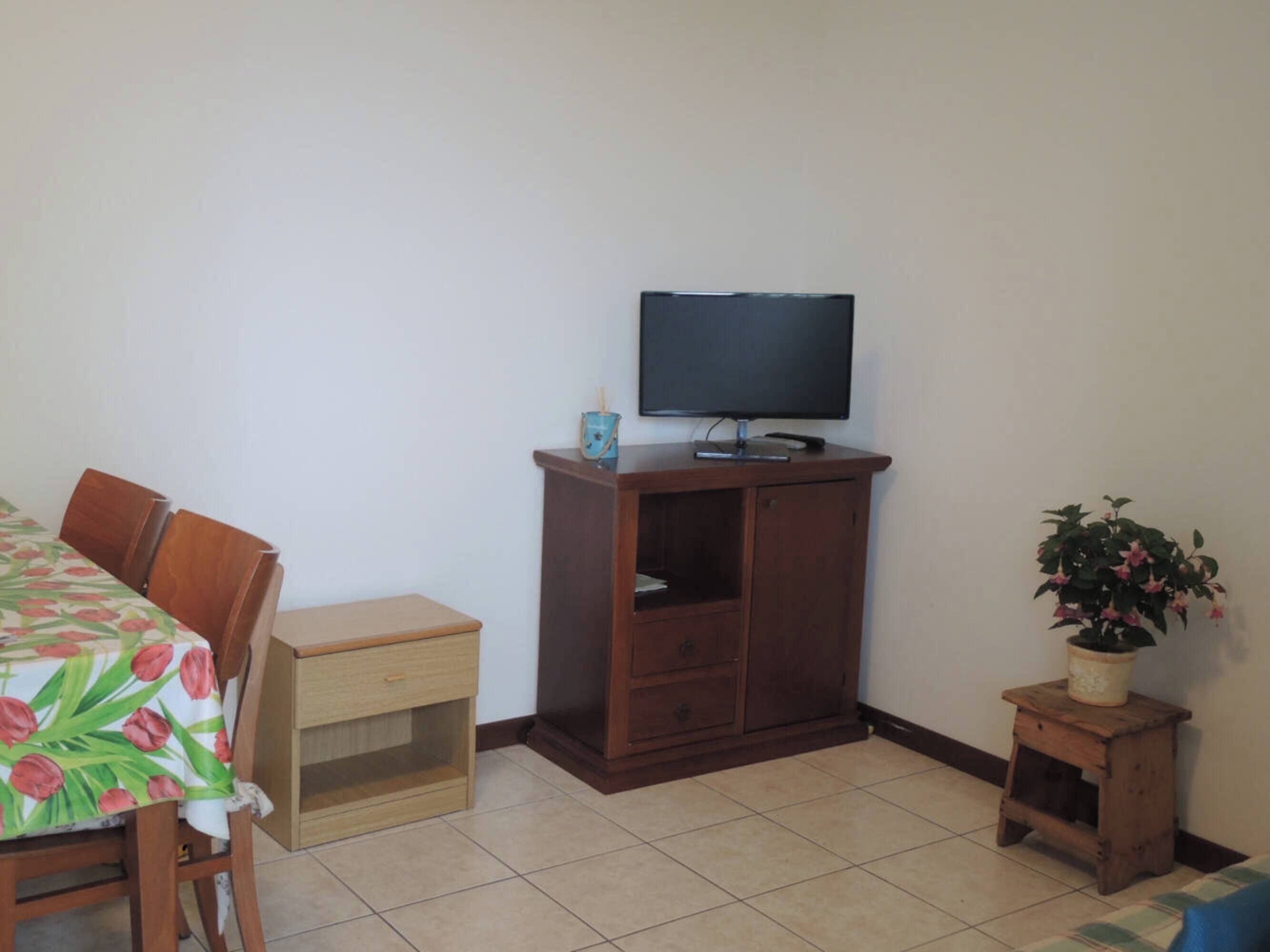 Ein kleines Zimmer mit einem Holzkasten, einem Fernseher darauf, einem kleinen Tisch mit einem Stuhl und einer Topfpflanze.
