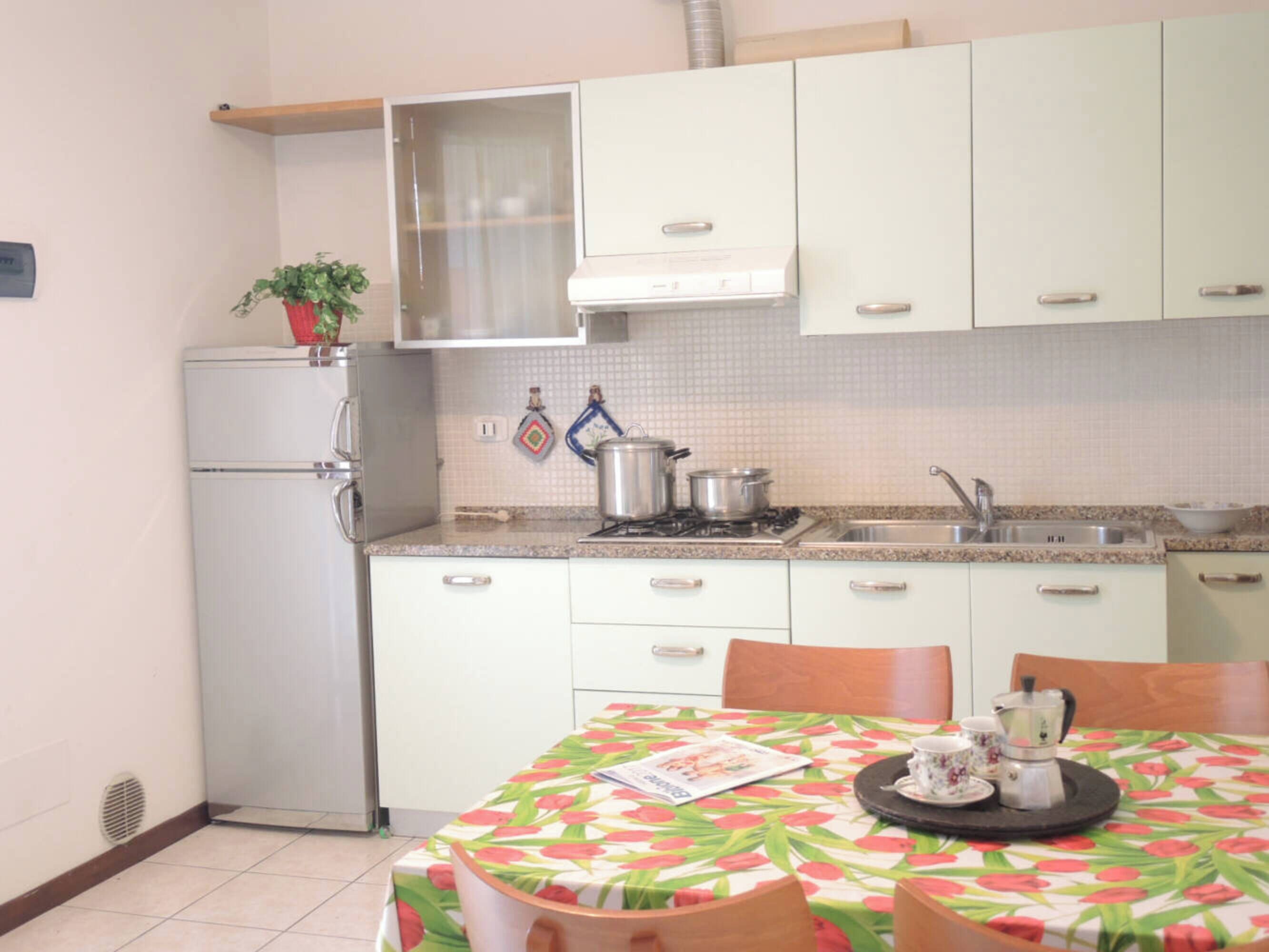 Une cuisine de taille réduite avec des meubles blancs, un réfrigérateur, une cuisinière et un évier. On y trouve une table recouverte d’une nappe à motifs floraux et un appareil à café.