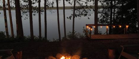 A campfire with a wooden bench and string lights, surrounded by trees and a lake.