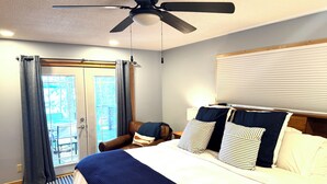 Ein Schlafzimmer mit Bett, Deckenventilator, Fenster mit Vorhängen, Nachttisch und Blick ins Freie.