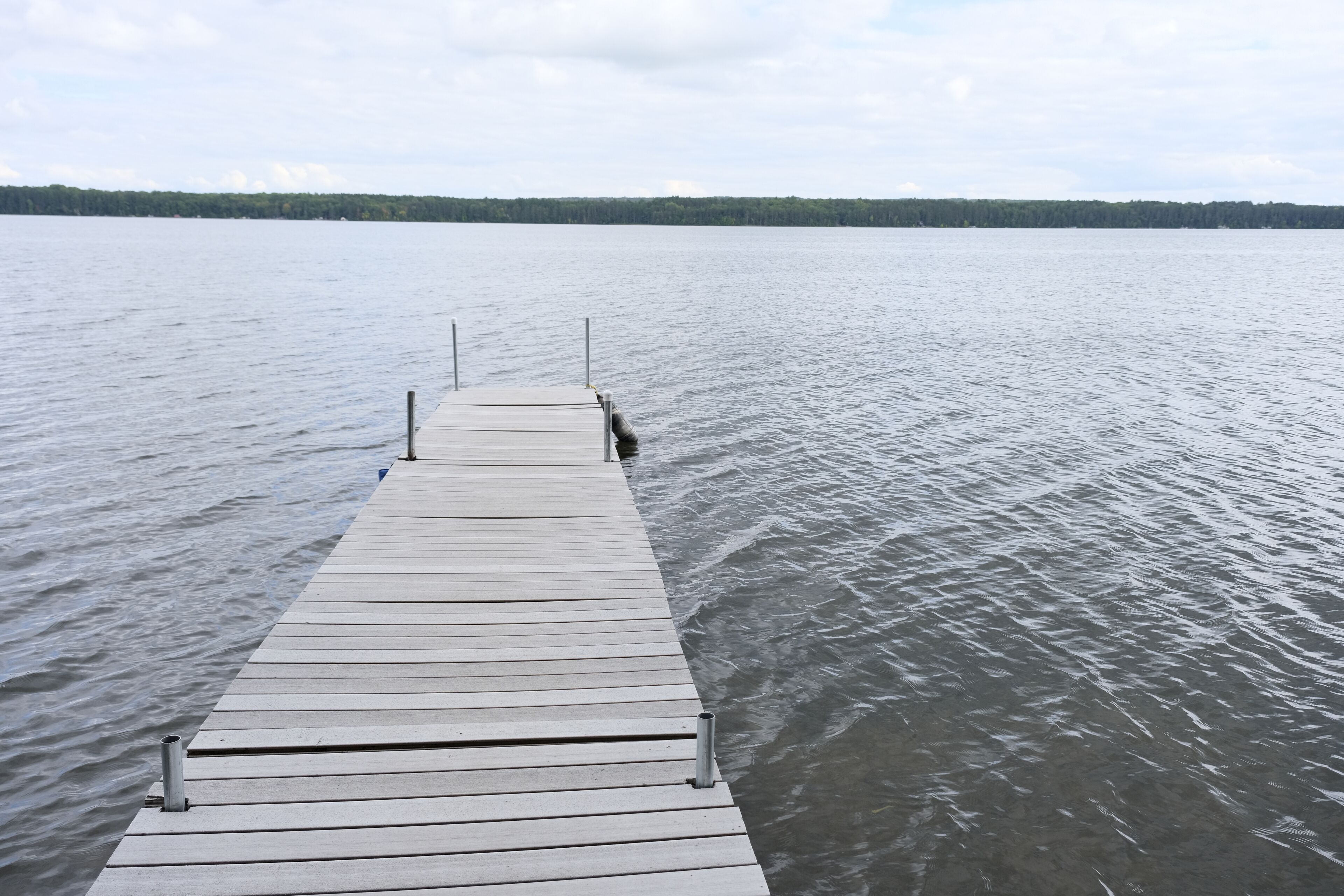 Un muelle de madera se adentra en un lago tranquilo con una orilla boscosa a lo lejos.