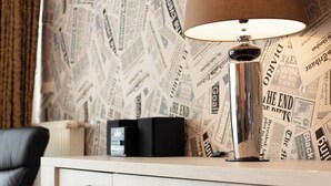 Una habitación con un mueble de madera claro con cajones, una silla negra y una lámpara con pantalla marrón. El papel tapiz de la pared detrás del mueble tiene un estampado de periódico.