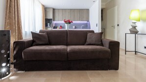 A modern living room with a brown sofa, a side table with a lamp, and a kitchen in the background.