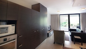 A modern kitchen with dark cabinetry, a microwave, and a built-in oven. There is a seating area with a sofa and a chair, and a large window providing a view of the outdoors. - Cosy Studio in Camps Bay (Cape Town)
