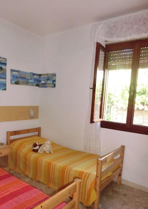 Une petite chambre avec un lit simple, une table de chevet, une fenêtre avec un rideau et des œuvres d’art accrochées au mur.
