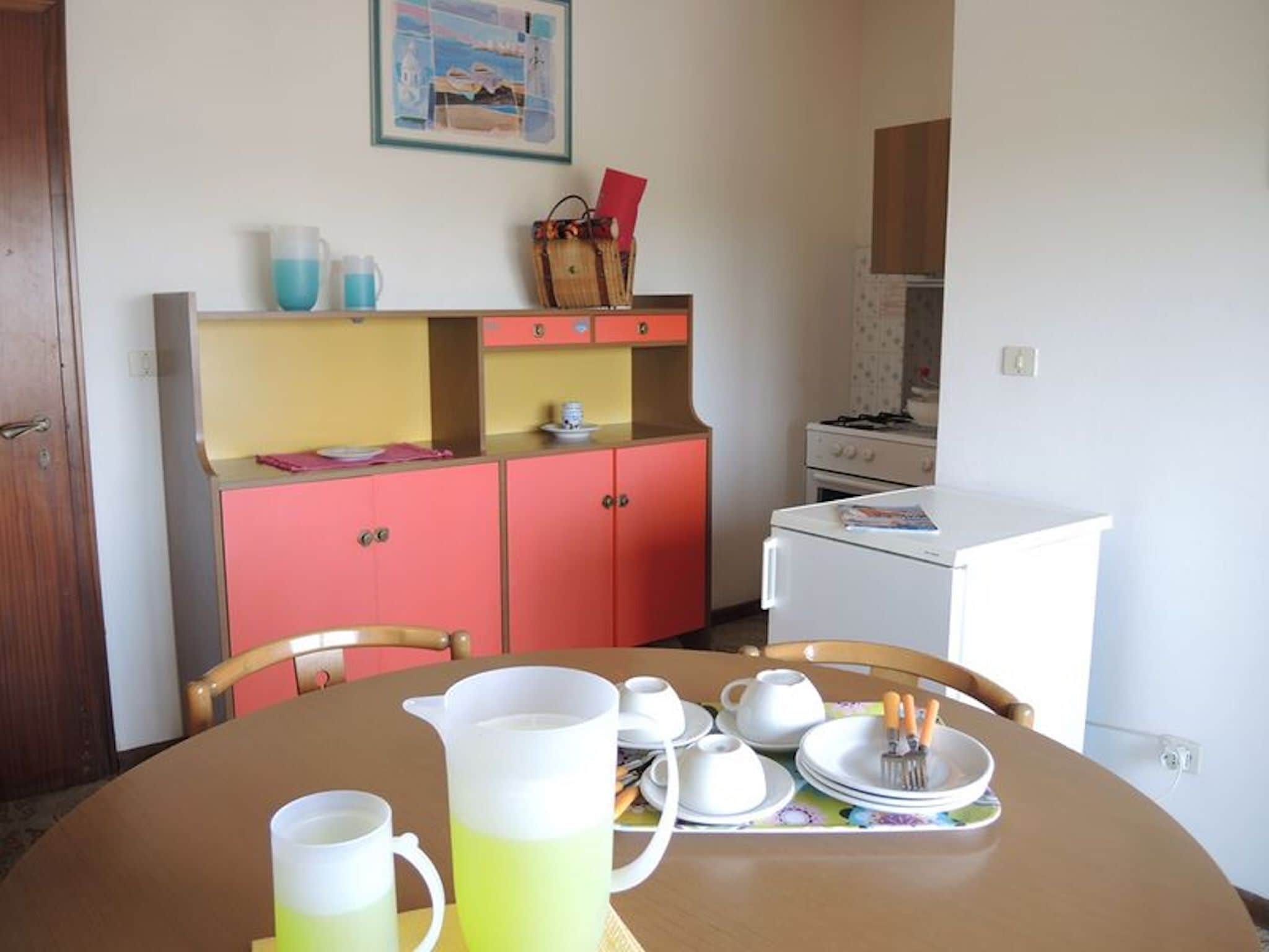 A compact kitchen with a white refrigerator, a small stove, and pink cabinets. There is a dining table with a pitcher of yellow liquid, cups, and plates.