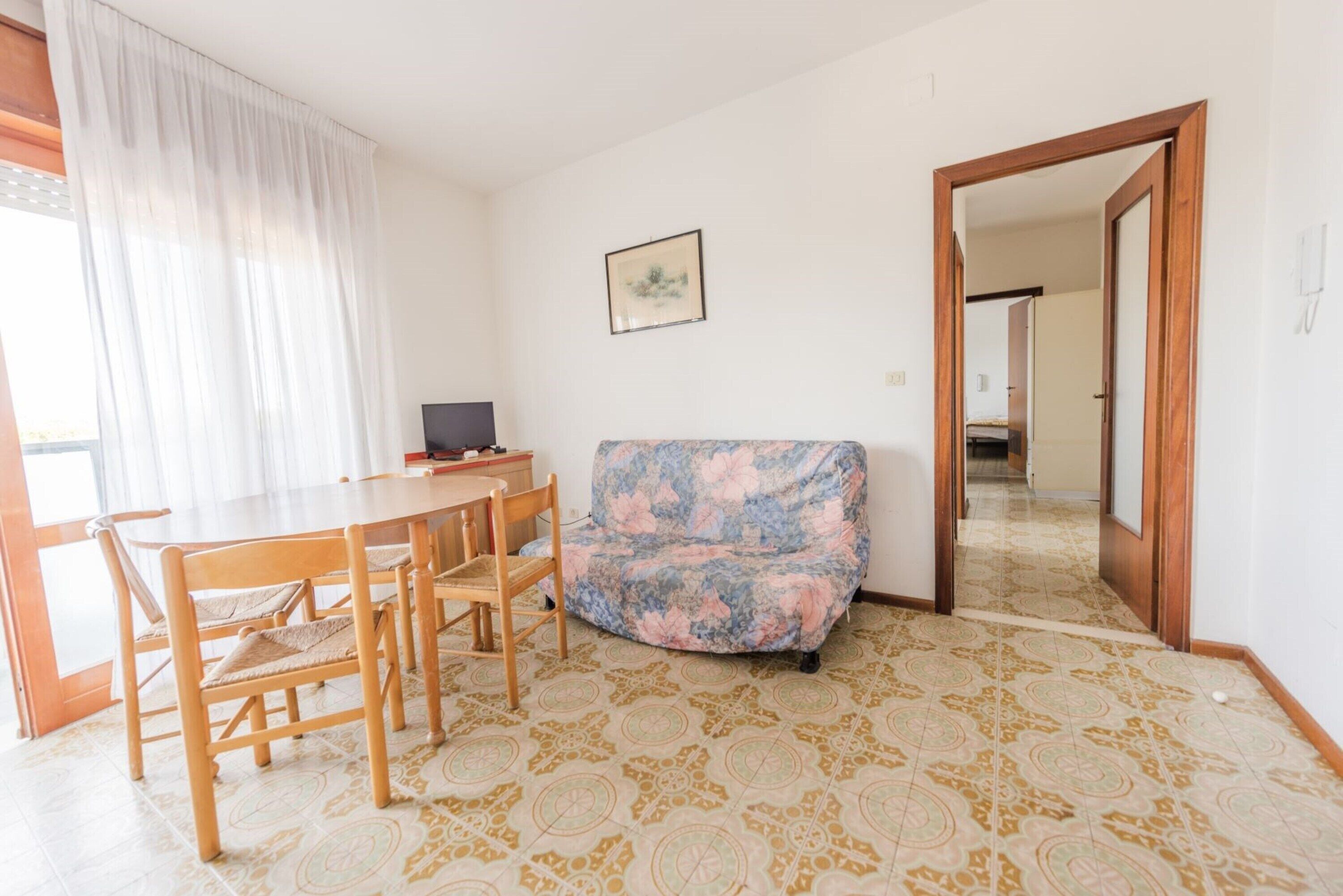 Ein Zimmer mit Esstisch, Stühlen, einer gepolsterten Couch, gemusterten Bodenfliesen, einem gerahmten Bild an der Wand und einer Tür zu einem weiteren Raum.
