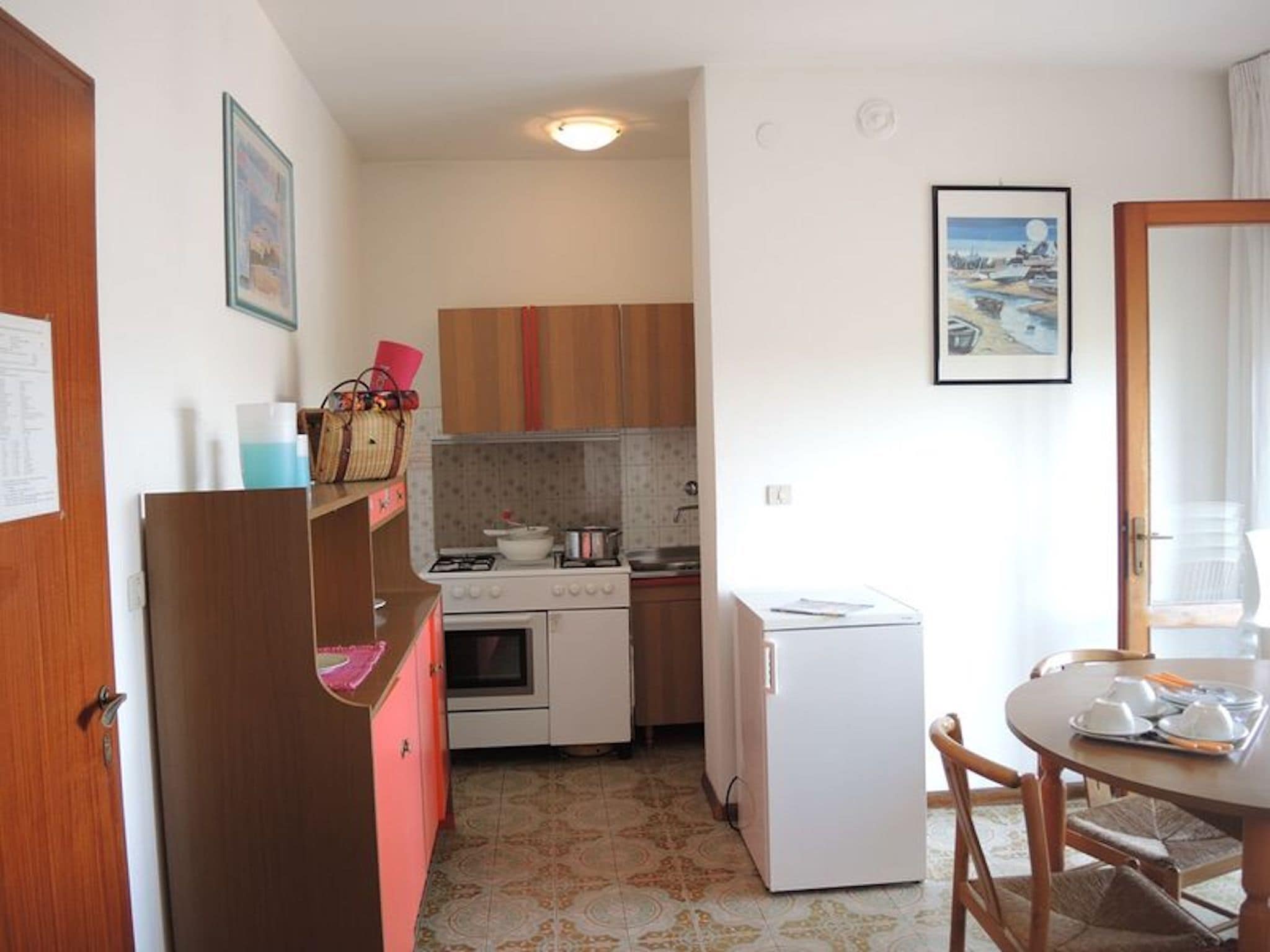 A compact kitchen with a refrigerator, oven, and microwave. There is a dining table with chairs and a small island. The kitchen has wooden cabinets and a tiled backsplash.