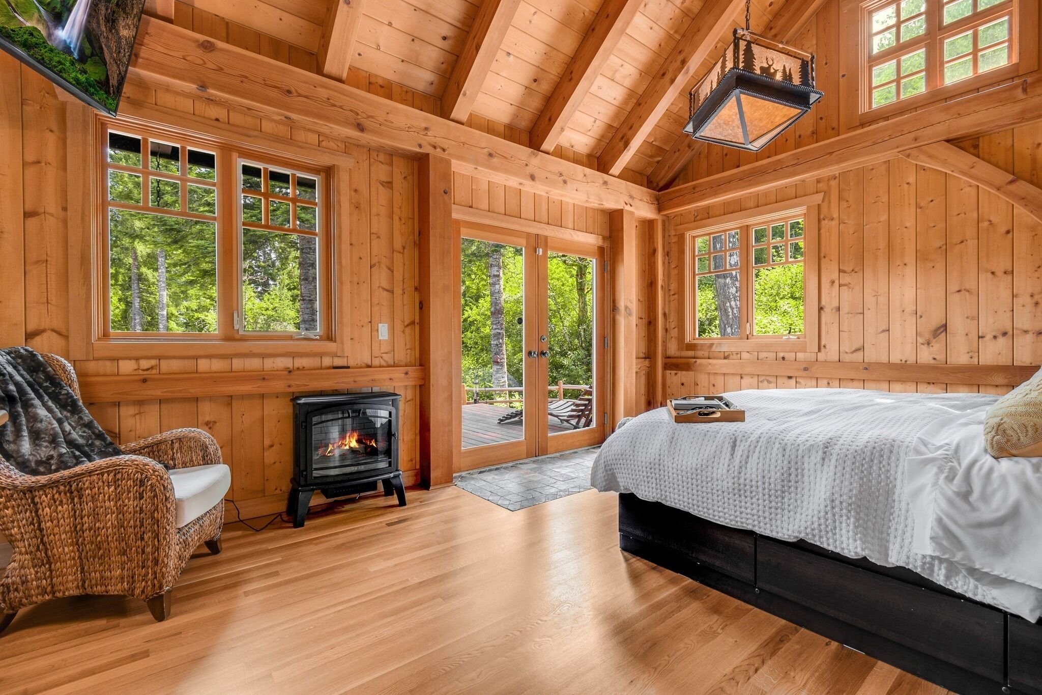L’intérieur d’un chalet en bois comprend un lit, un fauteuil en osier, une cheminée et de grandes fenêtres.