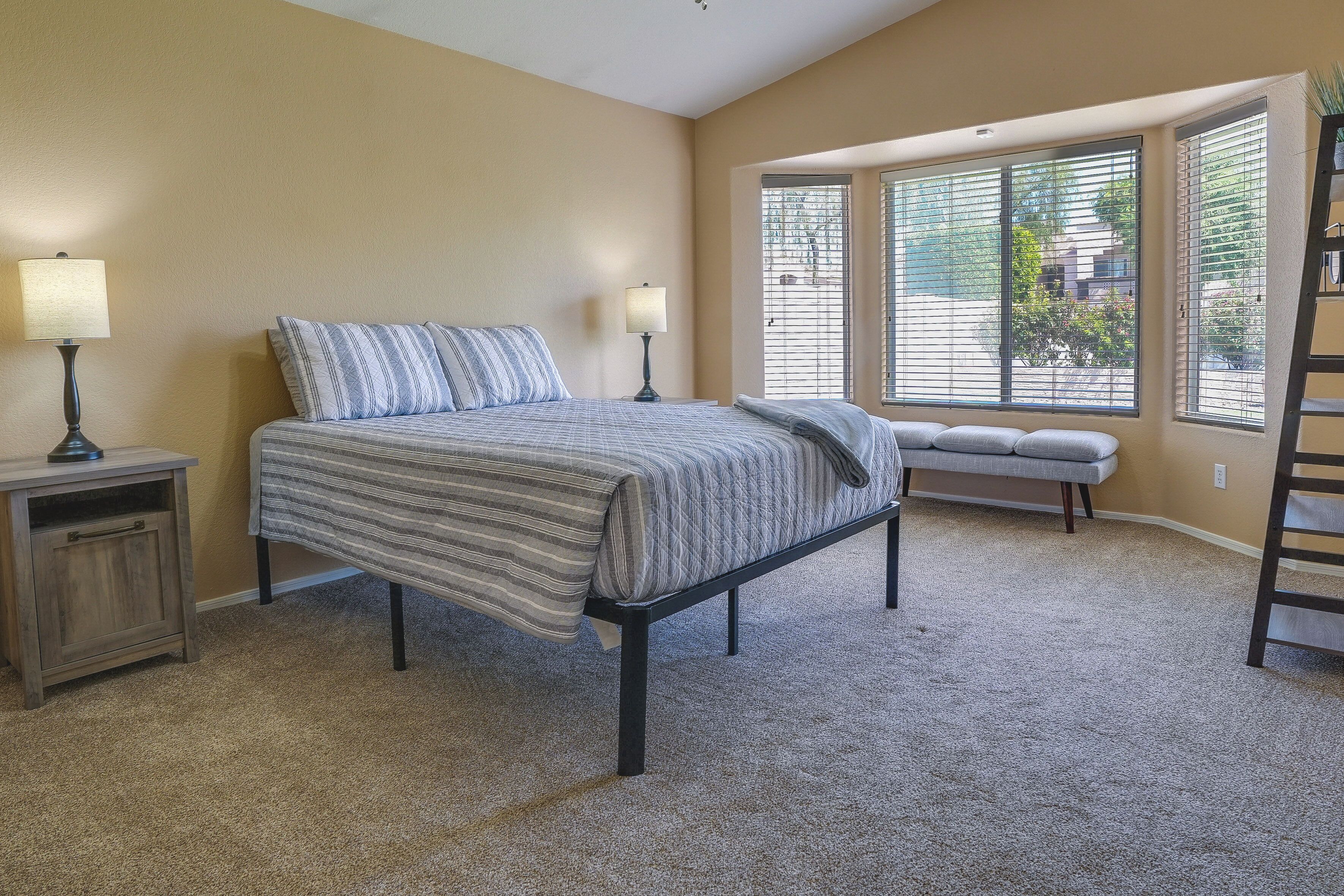 Une chambre à coucher avec un lit, deux tables de chevet avec des lampes, un banc et de grandes fenêtres équipées de stores.
