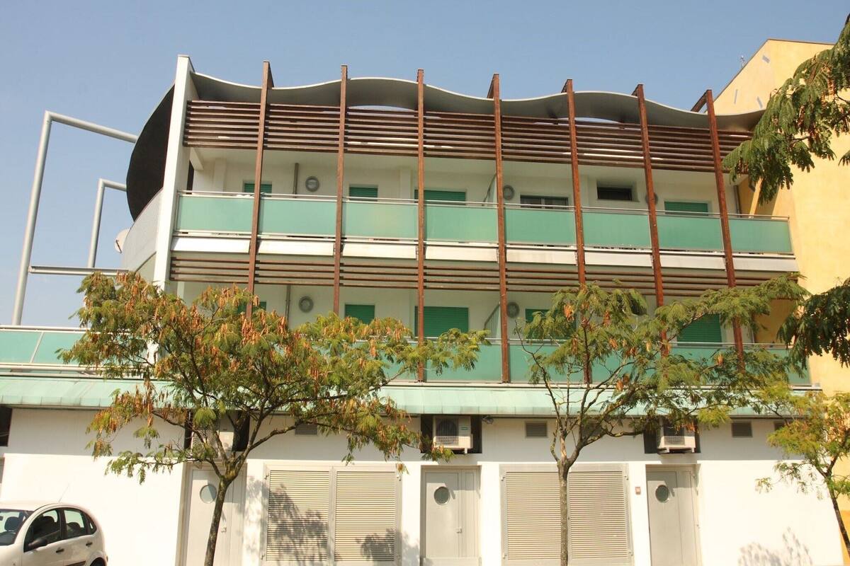 A multi-story building with a flat roof and a series of balconies. There are trees in front of the building, and a car parked on the left side.