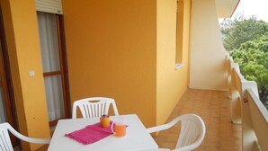 Ein kleiner Balkon mit Sitzgelegenheit im Freien, einem weißen Tisch und Stühlen, einer gelben Wand und Blick auf Grünflächen.