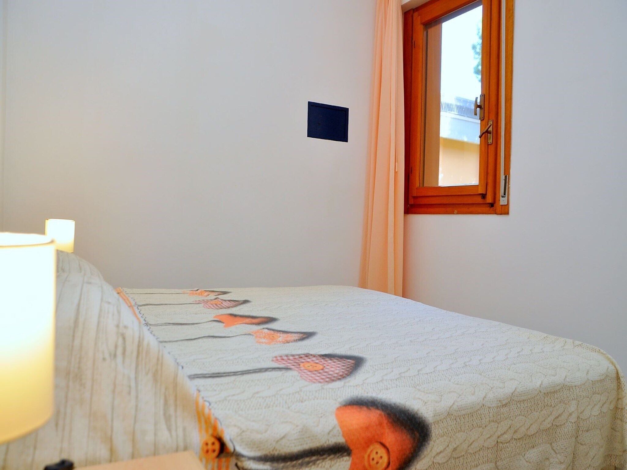 Un letto singolo con una coperta a motivi, una lampada da comodino, una finestra con tende e un piccolo oggetto fissato alla parete.
