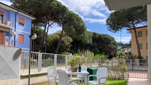 A patio area with white plastic chairs and a table, surrounded by a white fence, with buildings and trees in the background. - Modern Flat With Private Garden (Rosolina)