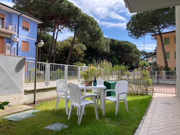 A patio area with white plastic chairs and a table, surrounded by a white fence, with buildings and trees in the background.