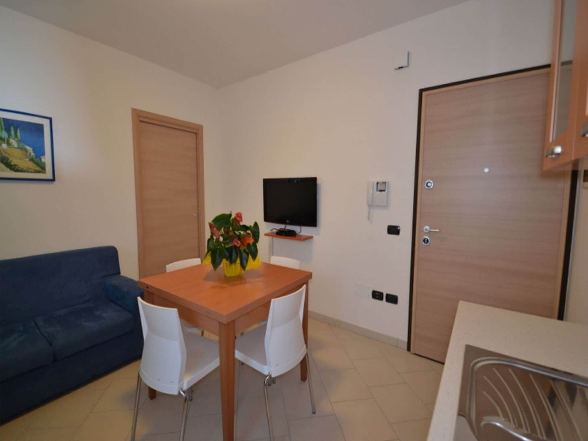 A small, well-lit room with a wooden dining table, white chairs, a blue sofa, a TV on a stand, and a wooden door.