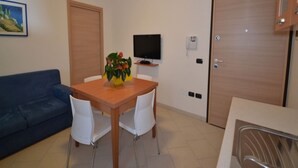 A small, well-lit room with a wooden dining table, white chairs, a blue sofa, a TV on a stand, and a wooden door. - Modern Flat With Private Garden (Rosolina)