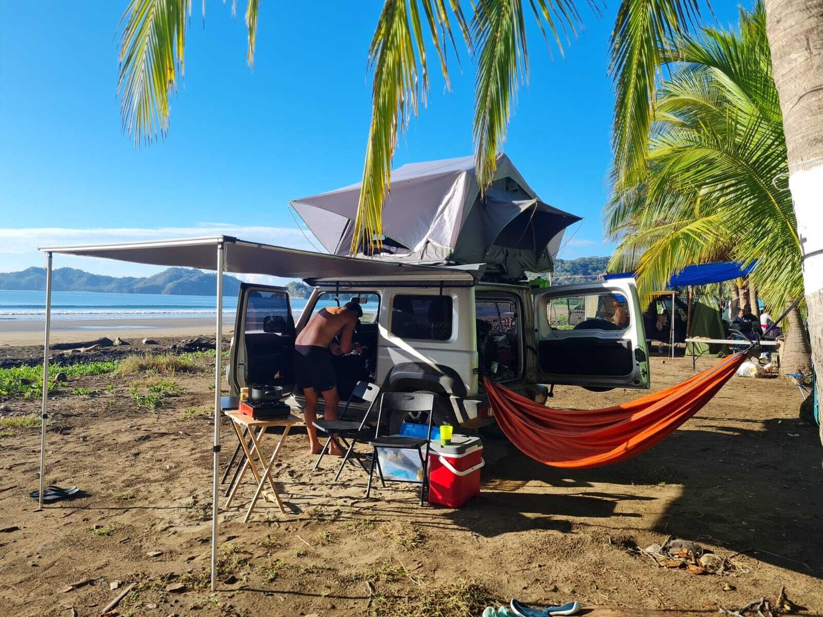 Un campament a la vora de la platja, amb un vehicle, una cadira d'hamaca i una nevera.