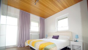 A bedroom with a wooden ceiling, a large bed with a colorful quilt, two bedside tables with lamps, and a window with curtains. - Ferienwohnung bis 3 Personen, auf 2 Ebenen, 80 qm, Balkon und Terrasse (Taching am See)