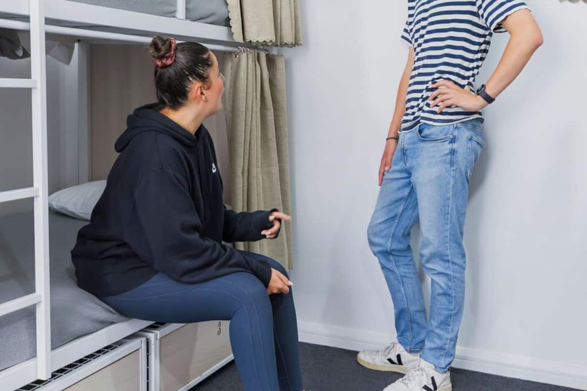 A bunk bed room with a person sitting on the lower bunk and another standing beside it.