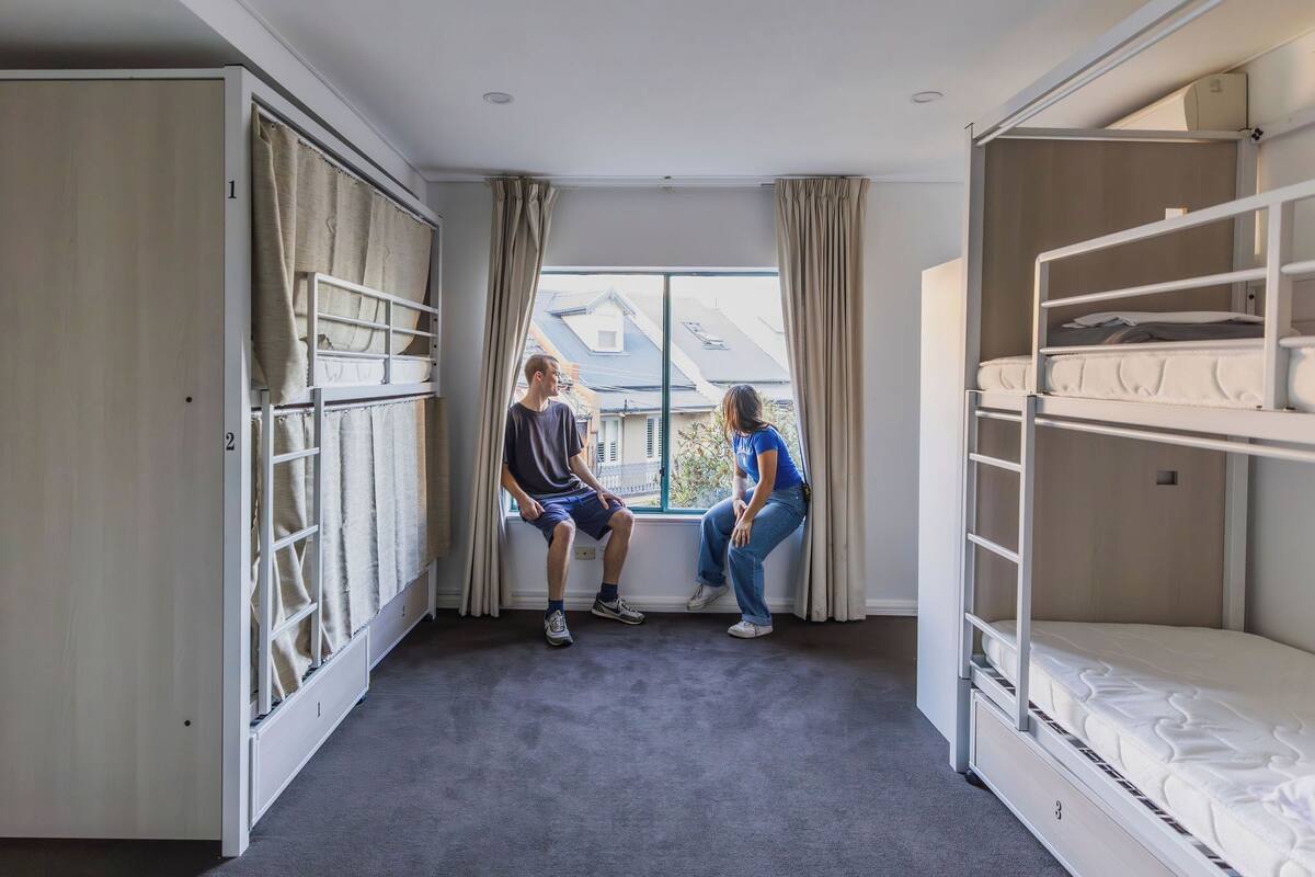 A room with bunk beds, a window with curtains, and a view of buildings outside.