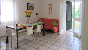 A dining area with a table, chairs, a vase with flowers, a sofa, and a door leading outside.