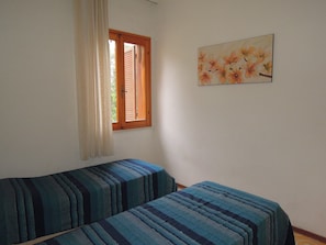 Una stanza con due letti singoli, una finestra con persiane di legno e un quadro floreale al muro.