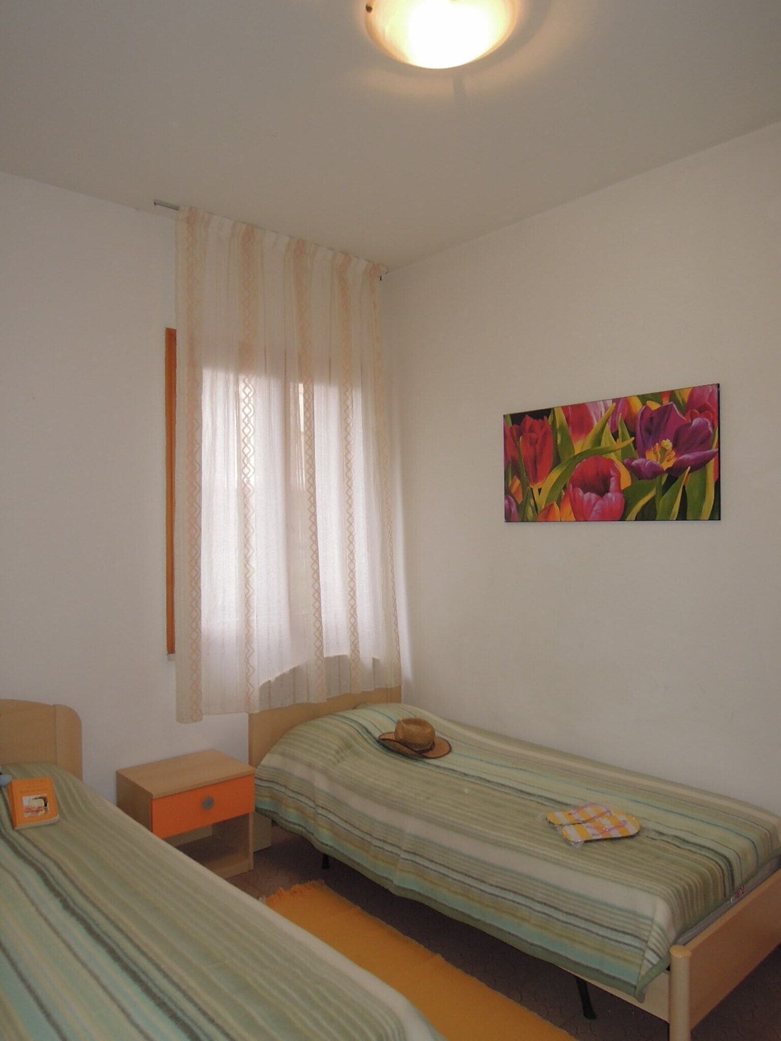 A bedroom with two single beds, a nightstand, a small table, and a window with sheer curtains.