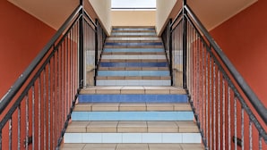 Eine Treppe mit abwechselnd blauen und beigen Stufen, schwarzen Geländern und einem Blick auf den Himmel durch ein Rundbogenfenster oben.