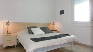 A neatly made bed with a white and gray duvet, two bedside tables with lamps, a small bedside table with a book and a plant, and a window with a view of the sky. - Precious Flat 100mt From the Beach - Beahost (San Michele al Tagliamento)