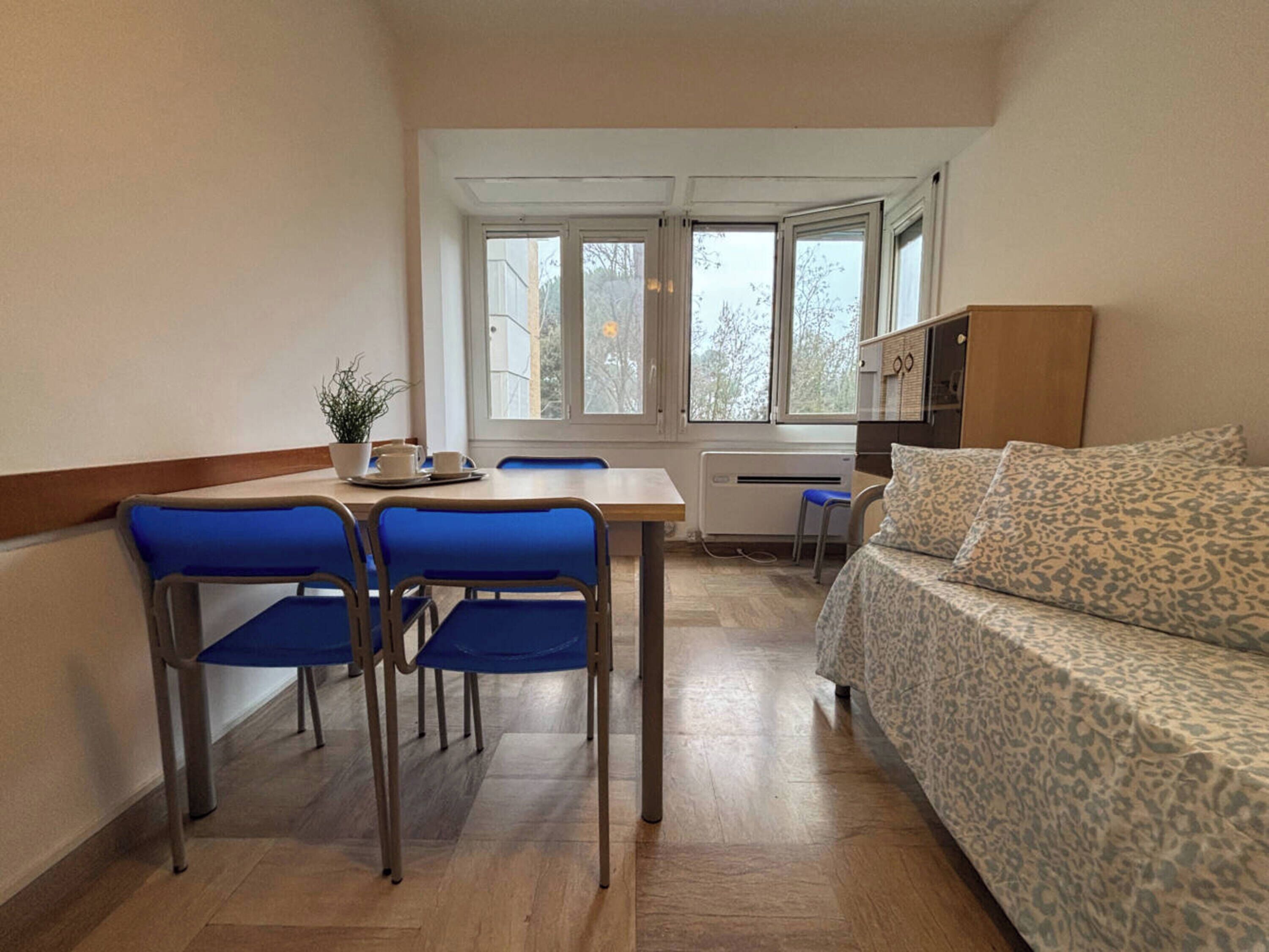 A compact room with a dining table, blue chairs, a sofa, and a window with a view of trees.