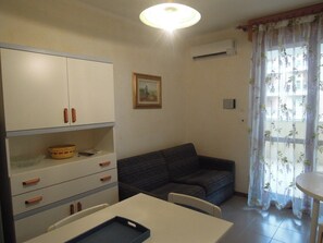A compact living space with a kitchenette, a sofa, a dining table, and a window with floral curtains. - Lovely Holiday Apartment With sea View (San Michele al Tagliamento)