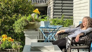 A person relaxing on a chair on a porch with a laptop, surrounded by potted plants and a blue table.