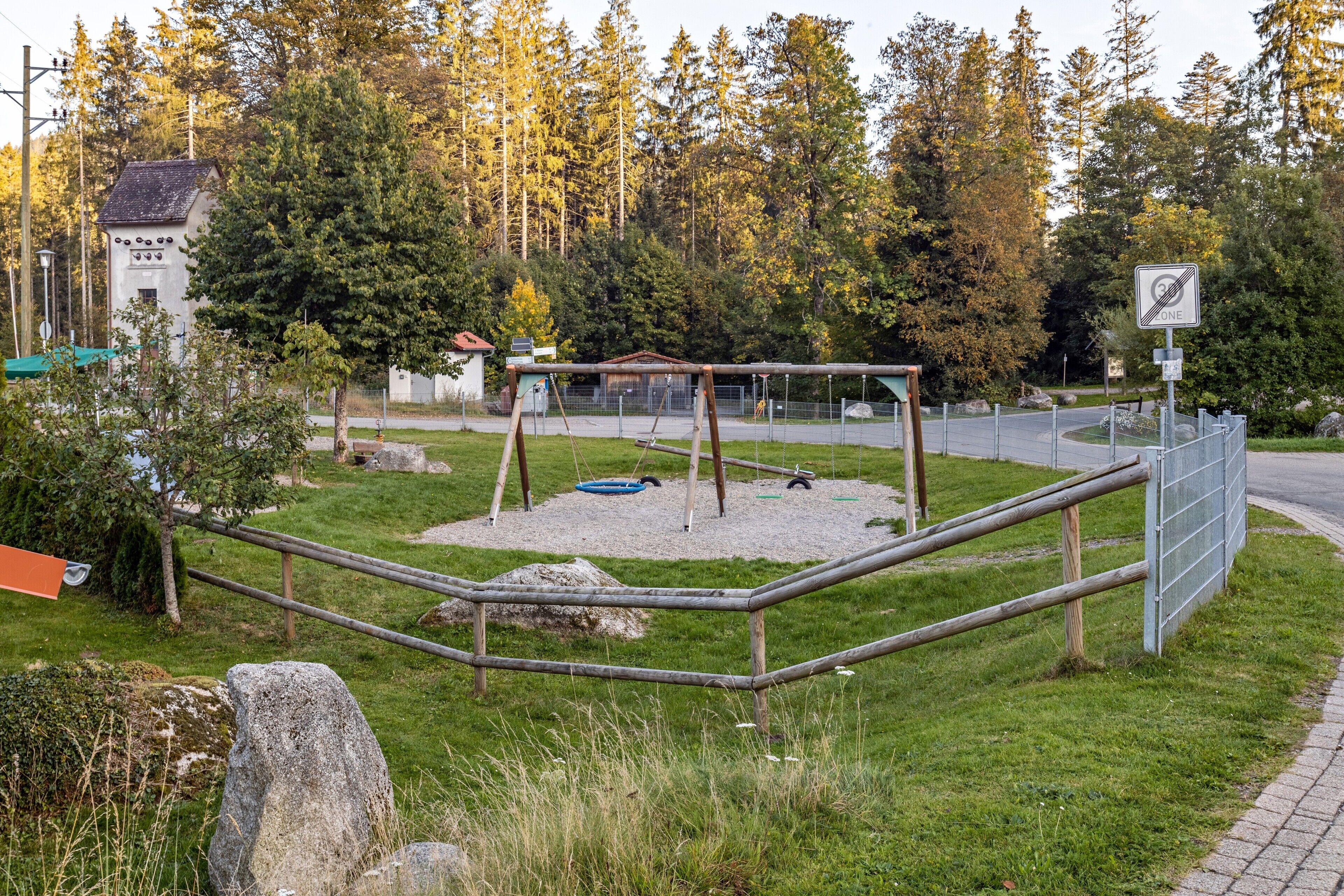 Un parque infantil con columpios y un área cercada, rodeado de árboles y un edificio con techo verde.