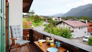A balcony with a wooden chair, a table with breakfast items, and a view of a village with houses and mountains. - Apartment 'Kramernest' with Mountain View, Balcony and Wi-Fi (Grainau)