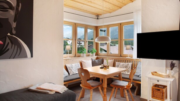 A cozy room with a wooden ceiling, a dining table with chairs, a bench, a TV, and large windows with a view of houses and mountains. - Apartment 'Kramernest' with Mountain View, Balcony and Wi-Fi (Grainau)