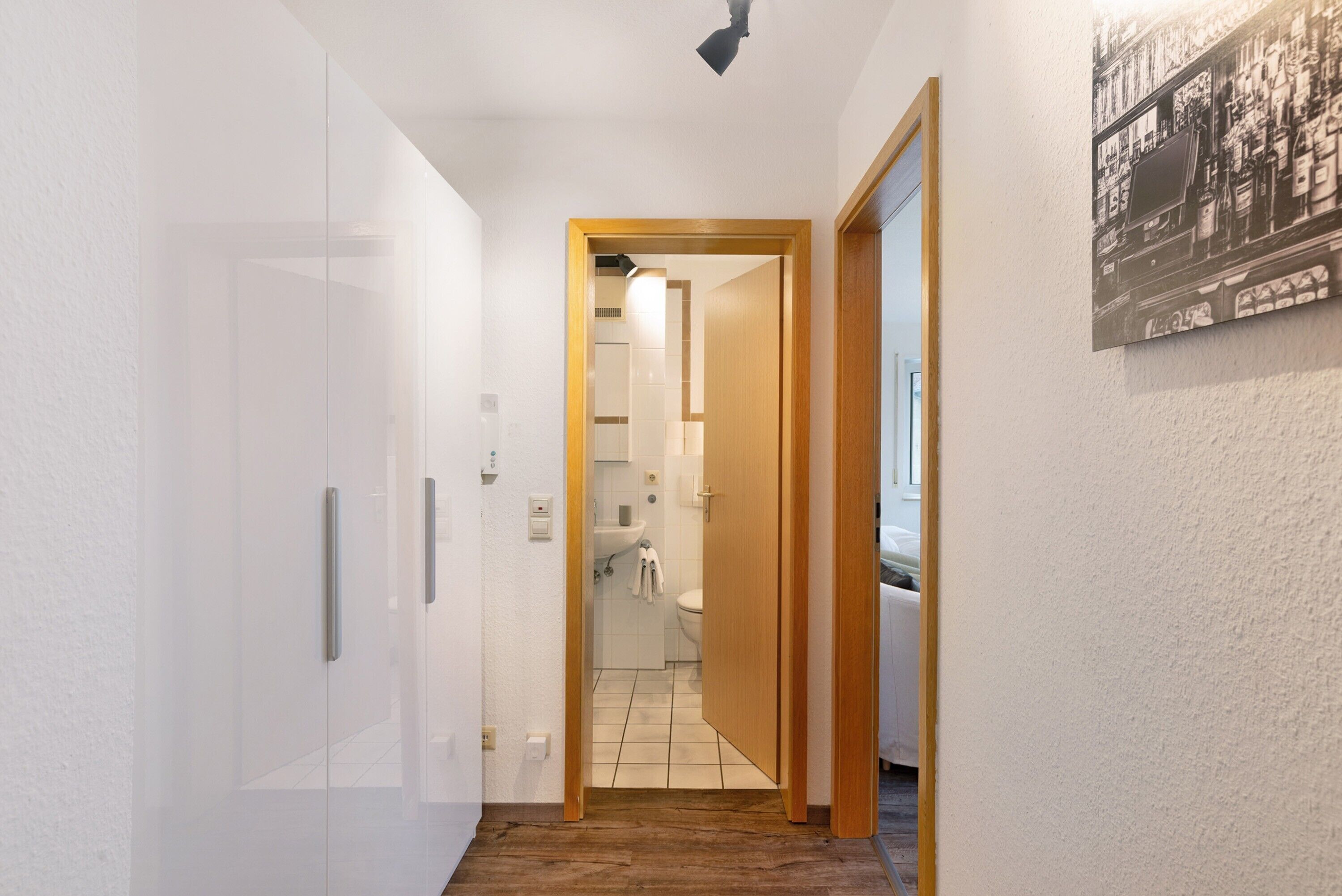Ein Flur mit einer Holztür zu einem Badezimmer, einer weißen Wand mit einem Plakat und einer Deckenleuchte.