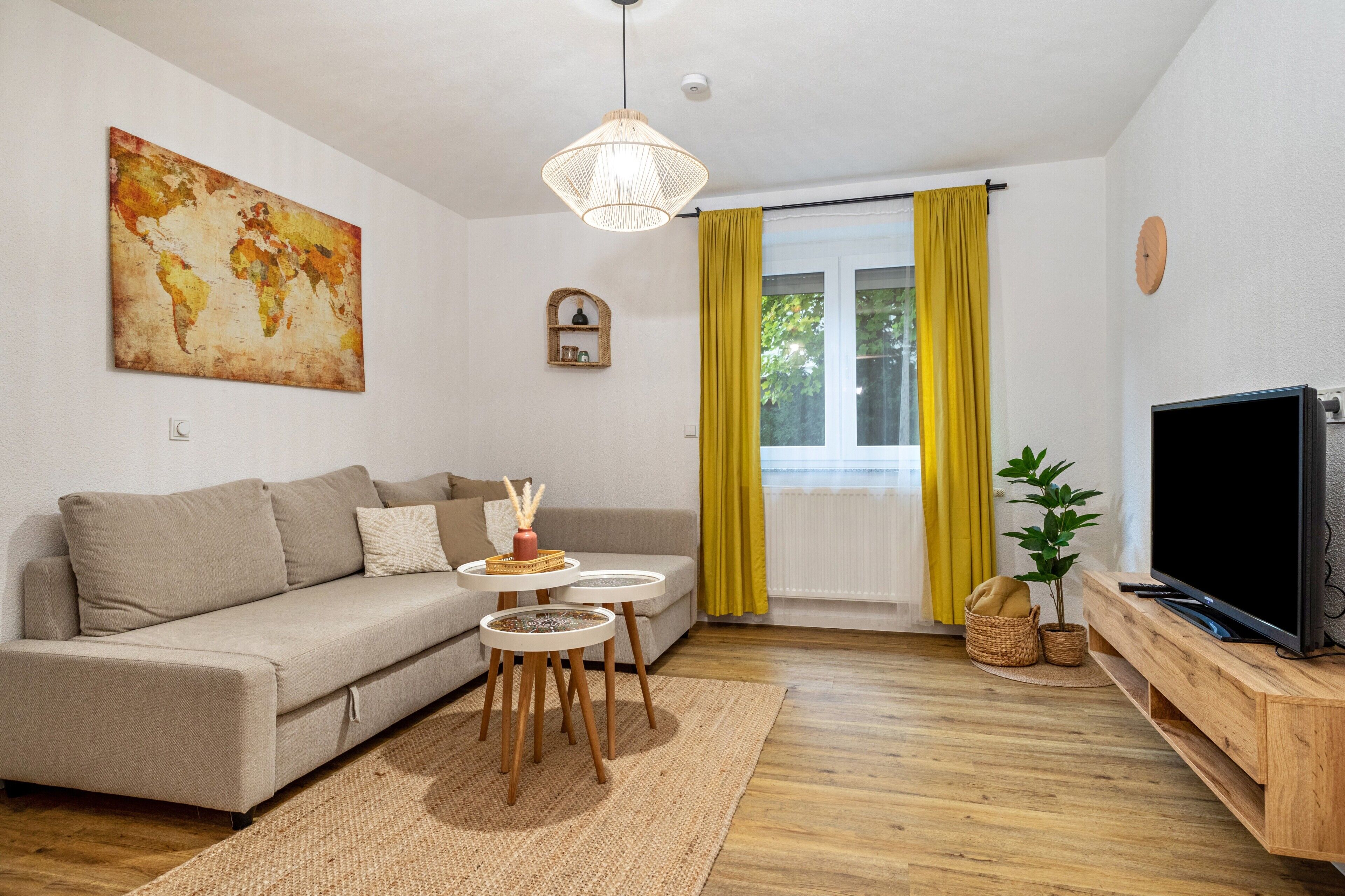 Ein Wohnzimmer mit einer Couch, einem Fernseher, einem Wandbild einer Weltkarte, einer Pendelleuchte und einem Fenster mit gelben Vorhängen.