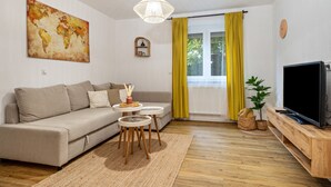 Una sala de estar con un sofá, un televisor, un cuadro de un mapa mundial, una lámpara colgante y una ventana con cortinas amarillas.
