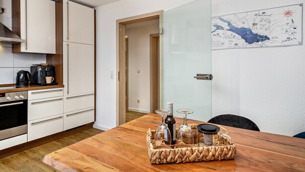 Una cocina moderna con armarios blancos, encimera de madera y una mesa de comedor con una cesta trenzada que contiene vasos y una botella.
