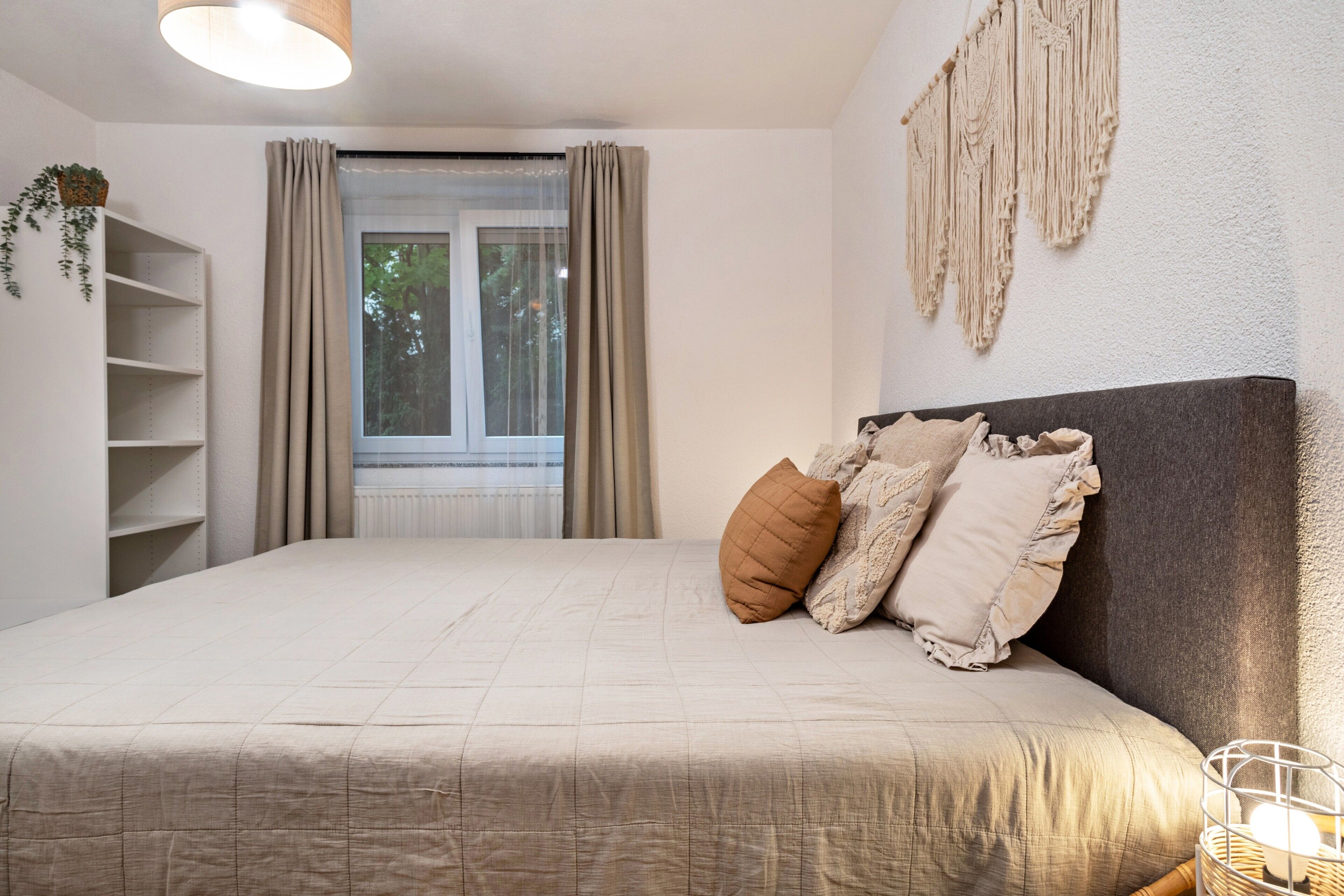 Ein Schlafzimmer mit Bett, Fenster mit Vorhängen, Wandbehagung aus Makramee und Nachttisch mit Lampe.