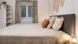 Un dormitorio con cama, ventana con cortinas, un macramé en la pared y una mesita de noche con lámpara.