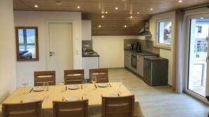 Een eetgedeelte met een tafel voor vier personen, houten stoelen, een keuken op de achtergrond en uitzicht door een raam.