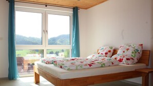 Een slaapkamer met een houten bed, een groot raam met gordijnen en uitzicht op een groen landschap.