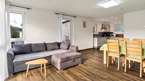 Ein modernes Wohnzimmer mit einer Couch, einem Hocker, einem Holztisch und Stühlen. Im Hintergrund ist eine Küche mit Geräten und einem Essbereich zu sehen.