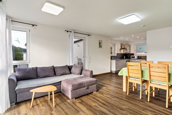 Un salon moderne avec un canapé, un pouf, une table en bois et des chaises. On aperçoit à l’arrière-plan une cuisine équipée et un coin repas.