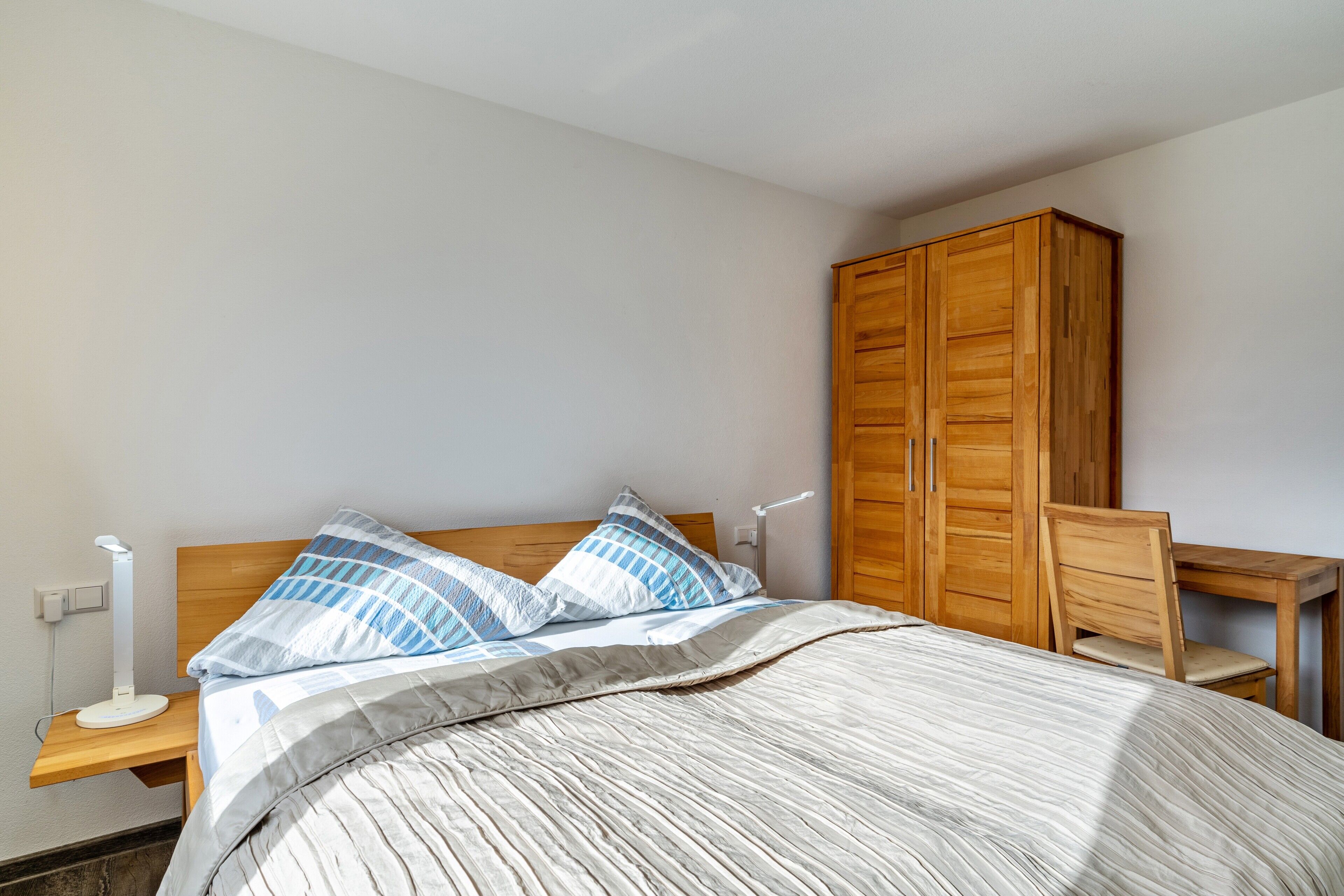 A bedroom with a bed, wooden wardrobe, bedside table, and a desk with a chair.