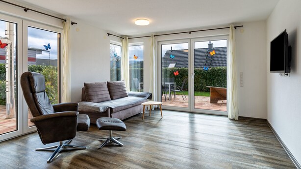 Amplia sala de estar con sofá, sillón y reposapiés, puertas correderas de cristal que dan a un patio, y un televisor de pantalla plana montado en la pared.