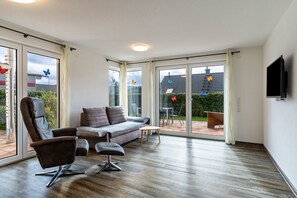 Ein großzügiger Wohnbereich mit Sofa, Sessel und Hocker, große Schiebeglasüren, die auf eine Terrasse führen, und einem wandmontierten Flachbildfernseher.