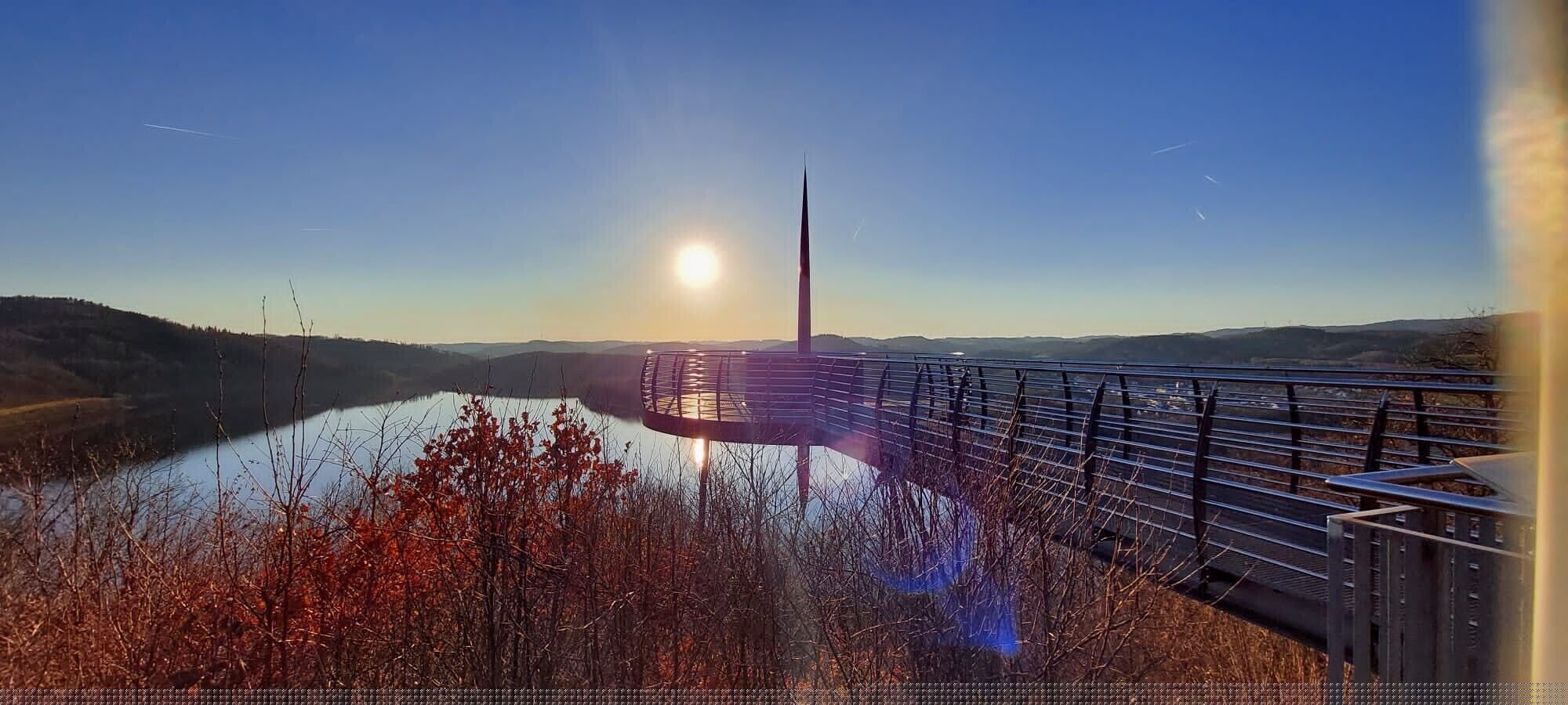 A bridge with a metal railing, a body of water, and hills in the background.